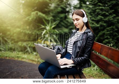 Smiling Student Sitting On Bench Listening Music And Holding Laptop In Park At University Preparing