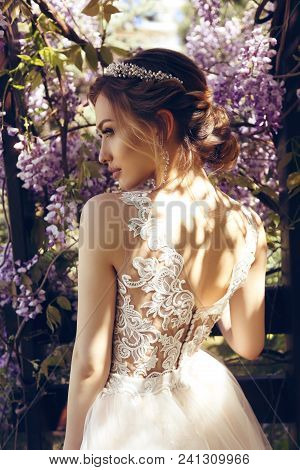 Bride  In Luxurious Wedding Dress And Diadema Posing Among Flowering Wisteria Trees