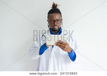 Serious Concentrated Young Black Male Doctor In Lab Coat Using Tablet And Looking At Camera. Frownin