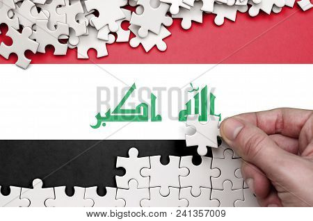 Iraq Flag  Is Depicted On A Table On Which The Human Hand Folds A Puzzle Of White Color.