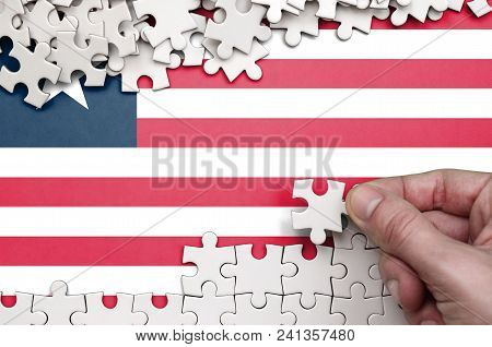 Liberia Flag  Is Depicted On A Table On Which The Human Hand Folds A Puzzle Of White Color.