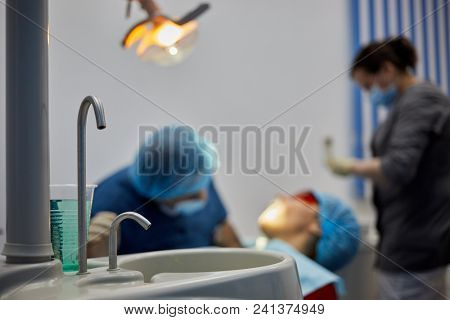 Dentist office with patient on dental chair, doctor and assistant, focus on taps and plastic cup with liquid.