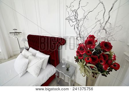 Bunch of artificial flowers, branches, berries in vase in bedroom with red bed.