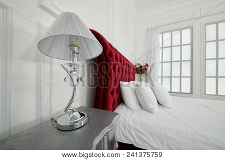 Lamp on bed-table and large red bed in white bedroom with latticed window.