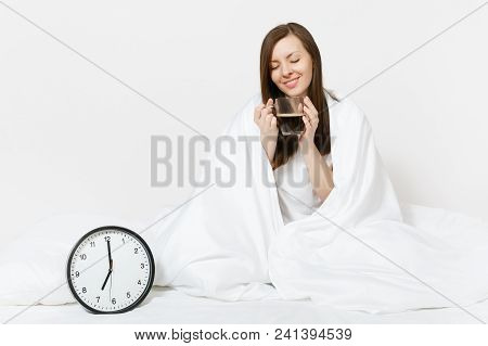 Happy Woman Sitting In Bed With Round Clock, White Sheet, Pillow, Wrapping In Blanket On White Backg