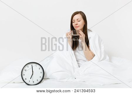 Happy Woman Sitting In Bed With Round Clock, White Sheet, Pillow, Wrapping In Blanket On White Backg