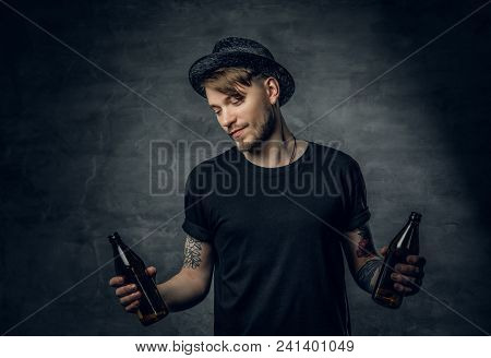 Handsome Bearded Male With Tattooed Arms, Dressed In A Black T Shirt And Top Hat Holds Craft Bottled