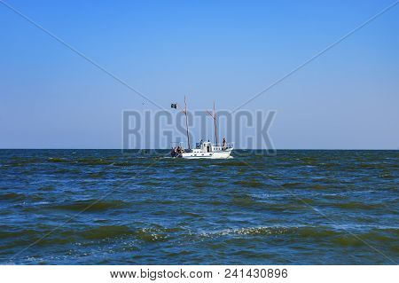 Small Boat With Tourists On Board Touring The Black Sea. A Ship With A Pirate Flag Sails On Blue Wav