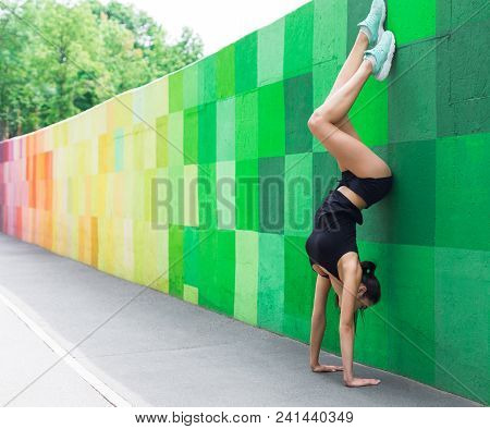 Young Woman Doing Handstand Exercise Against Green Wall On City Street. Sporty Girl Practicing Pilat
