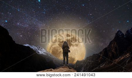 A Man With Backpack Standing On Mountain Peak At Night, And Silhouette Mountain With Bright Full Moo