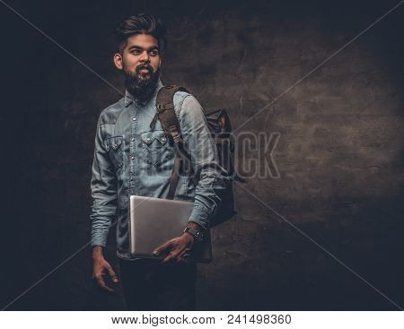 The Indian Male Student Holds Laptop And Backpack Over Grey Studio Background.