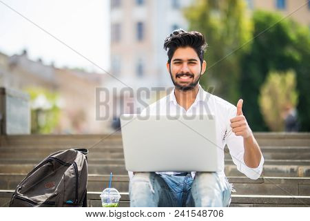 Happy Indian Student Sitting On The Stairs Showing Thumb Up Working On Laptop, In The University Cam