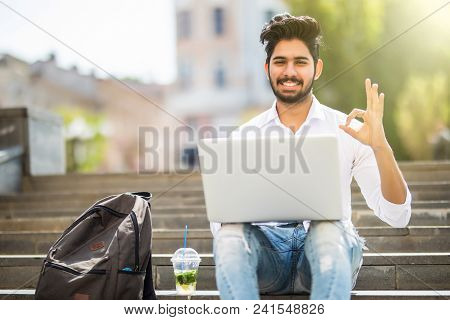 Happy Indian Student Sitting On The Stairs Showing Okay Sign Working On Laptop, In The University Ca