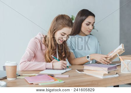 Multiethnic Girls Studying Together And Writing In Copybook And Reading Book At Table