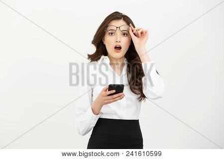 Business Concept: Surprised Youngwoman Holding Mobile Phone And Staring At It While Standing Against