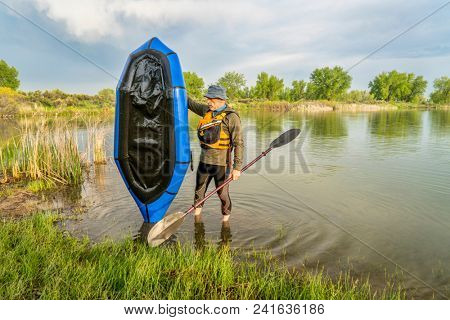 a senior male with a blue pacraft (one-person light raft used for expedition or adventure racing) on a lake shore in spring scenery