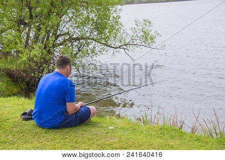 A Man With A Fishing Rod Fishing In The Pond, On A Sunny Lake Sitting On The Shore