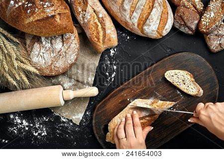 Woman Hands Cutting A Loaf Of Bread On Rustic Wooden Board, With Wheat Ears And Knife, Top View. Sti