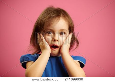Scared Little Adorable Girl With Wide Open Mouth, Big Bright Brown Eyes And Hands On Cheeks Dressed 