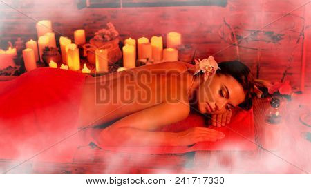 Massage of woman in spa salon. Girl on candles aglow background in steam room. Luxary interior in oriental therapy salon. Photo effect with red toning.
