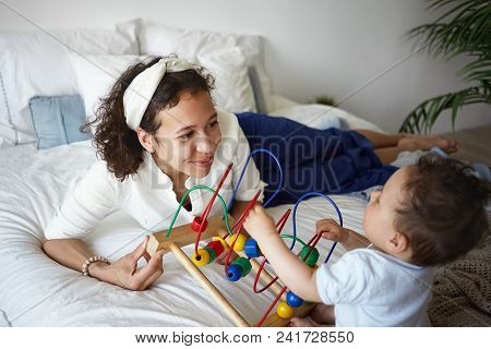 Picture Of Stylish Young Hispanic Female Smiling Joyfully, Teaching Her Baby Infant How To Play New 