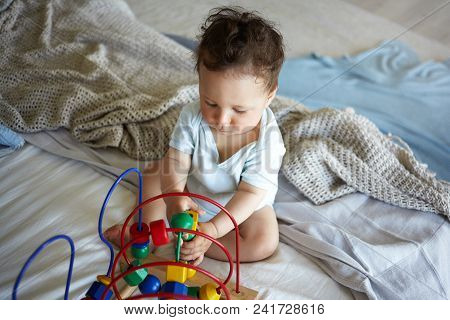 Close Up Shot Of Serious Boy Toddler Sitting On Undone Bed, Having Concentrated Focused Look, Playin