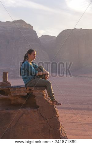 The Girl-traveler Sits On A Stone Ledge Against The Backdrop Of The Mountains In The Wadi Wadi Deser