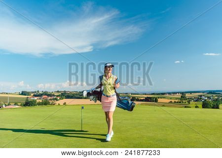 Full length rear view of an active woman carrying a blue stand bag, while walking on the grass towards a training area for professional golf in a sunny day
