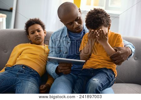 Indulging A Child. Pleasant Loving Father Offering A Tablet To His Crying Little Son Sitting On His 