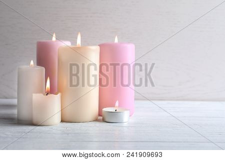 Different Wax Candles Burning On Table Against Light Background