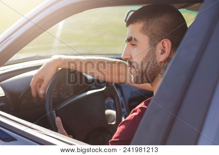 Sideways Shot Of Handsome Young Unshaven Male Sits At Wheel In His New Car, Has Thoughtful And Conce