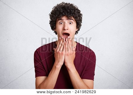 Photo Of Astonished Male Teenager, Keeps Hands On Chin, Mouth Widely Opened, Being Stunned By Bad Ne
