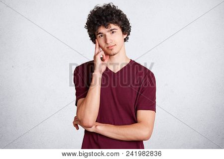 Waist Up Portrait Of Handsome Male Keeps Hand Near Temple, Has Pensive And Satified Expression, Wear