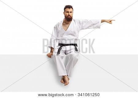 Man in karate kimono with black belt sitting on a blank panel and pointing to one side isolated on white background