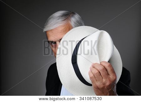 Closeup of a middle aged man wearing a tuxedo and a Panama Hat in his hand as he peeks around the hat. Horizontal over a gray background.