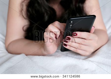 Woman With Long Hair Lying On A Bed And Using Smartphone. Mobile Phone In Female Hands, Concept Of O