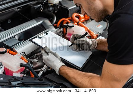 Cropped View Of Mechanic Writing On Clipboard While Inspecting Car Engine Compartment