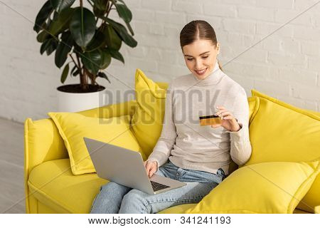 Smiling Girl Looking At Credit Card And Using Laptop On Sofa