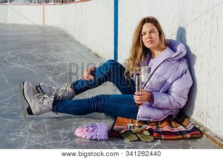 Lovely Young Woman Relaxing After Riding Ice Skates And Drinking Hot Drink From Termo Pot On The Ice