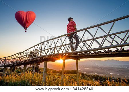 Silhouetted Of Asian Man Standing On Bamboo Bridge With Red Hot Air Balloon In The Shape Of A Heart 