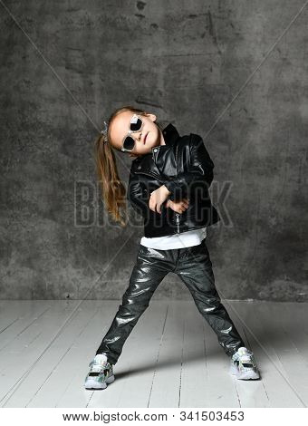 Cheerful Frolic Kid Girl In Leather Jacket, Stylish Jeans And Sunglasses Is Having Fun Playing Danci