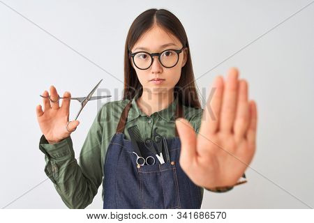 Chinese hairdresser woman wearing glasses holding scissors over isolated white background with open hand doing stop sign with serious and confident expression, defense gesture