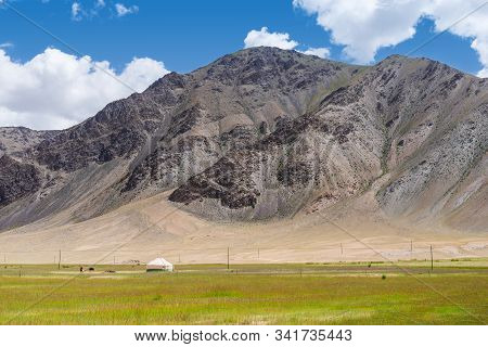 Vibrant Green Pasture With Yurt And Farmer At The Foot Of Barren Rocky Mountains Under Cloudy Blue S