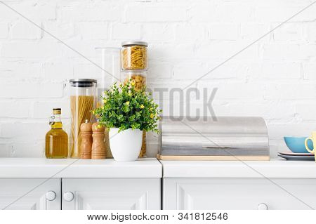 Modern White Kitchen Interior With Breadbox, Pasta, Oil And Green Plant Near Brick Wall