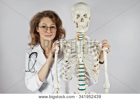 Woman In Medical Gown. Doctor With Skeleton Layout. Girl Medic In Glasses Isolated On A White Backgr