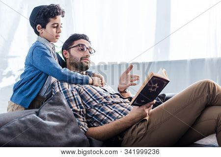 Jewish Father Pointing With Hand At Tanakh And Talking With Son In Apartment