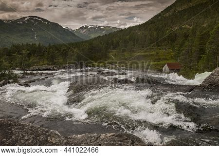 Raw Waterfall With Small House And Mountains With Snow As Background Near Balestrand