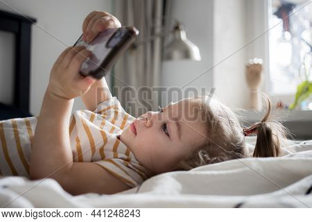 Little Toddle Girl Playing With Smartphone Laying On The Bed At Home. Caucasian Toddler Girl. High Q