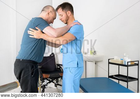 Male Physiotherapist Helping A Patient With A Disability Who Uses A Wheelchair, To Get Up At Rehabil
