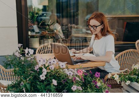 Young Happy European Woman Sitting At Cafe Table Outdoors With Laptop And Cup Of Coffee, Smiling Red